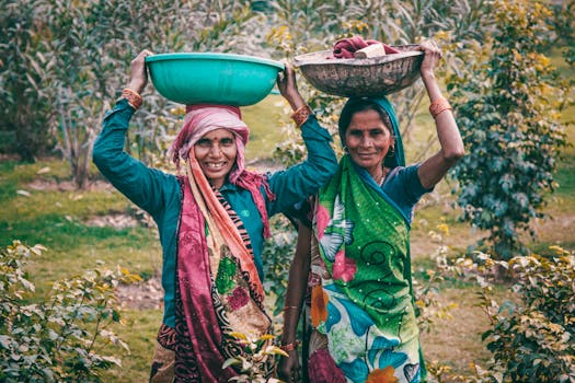 Two women wearing colorful traditional dresses smile outdoors, carrying baskets on their heads.